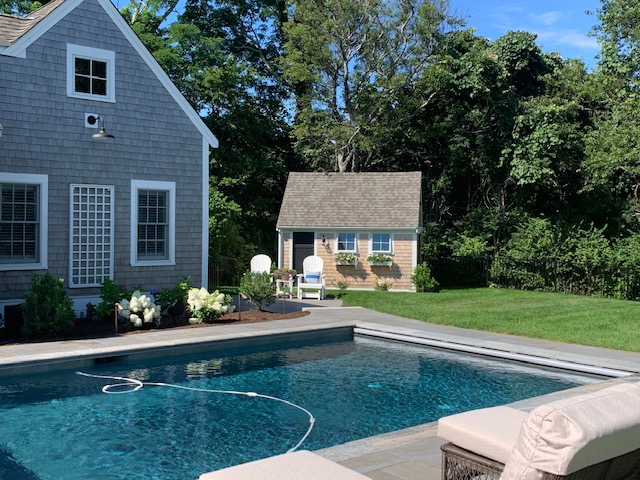 A pool with hardscaping and a cute shed on a lawn on Cape Cod